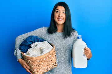 Beautiful asian young woman holding laundry basket and detergent bottle smiling looking to the side...