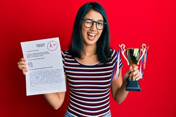 Beautiful asian young woman showing a passed exam holding trophy winking looking at the camera with...