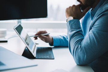 Crop businessman using stylus while browsing tablet placed on keyboard