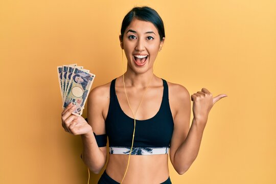 Beautiful asian young sport woman holding 5000 japanese yen banknotes pointing thumb up to the side smiling happy with open mouth