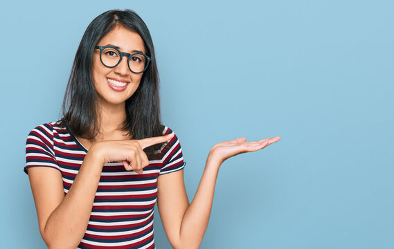 Beautiful asian young woman wearing casual clothes and glasses amazed and smiling to the camera while presenting with hand and pointing with finger.