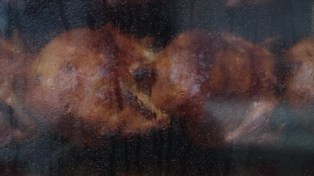 Close-up shot of a steamy rotisserie machine window with water drops. Grilled roasted whole chickens with tasty golden-yellow roasted skin on a turning spit in a row. Restaurant, butcher shop roaster.