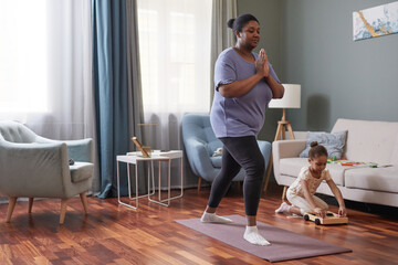 Fototapeta na wymiar Full length portrait of African-American woman doing yoga at home with little girl in background, copy space