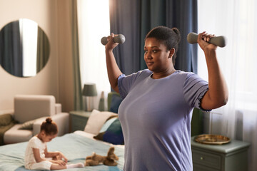 Waist up portrait of African-American woman working out with dumbbells at home with little girl in background, copy space