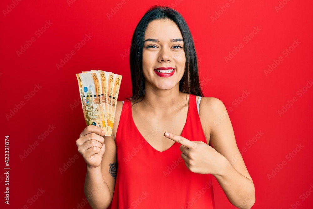 Poster Young hispanic girl holding 500 philippine peso banknotes smiling happy pointing with hand and finger