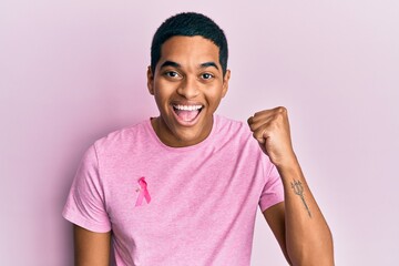 Young handsome hispanic man wearing pink cancer ribbon on shirt screaming proud, celebrating...