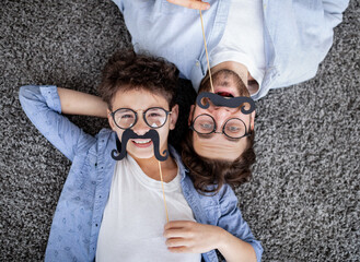 Funtime together. Curly father and son playing with fake moustache on sticks, lying on floor, top...