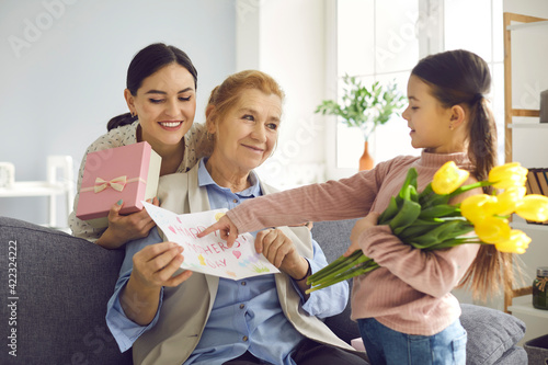 Surprise for the dearest. Little daughter and her adult mother congratulate the grandmother on Mother's Day. Girl points to a handmade card held by an older woman. Family, women and gifts concept.