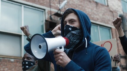 Caucasian young handsome man in hood and scarf on face screaming in megaphone at protest for human...