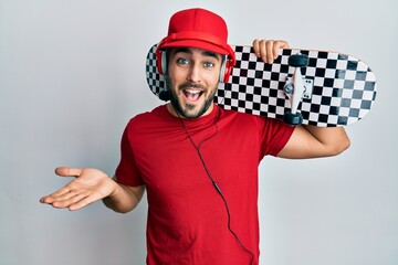 Young hispanic man using headphones holding skate celebrating achievement with happy smile and winner expression with raised hand