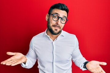 Young hispanic businessman wearing shirt and glasses clueless and confused expression with arms and...
