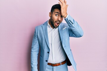 Young hispanic businessman wearing business jacket surprised with hand on head for mistake,...