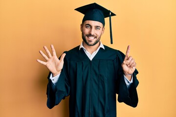 Young hispanic man wearing graduation cap and ceremony robe showing and pointing up with fingers number six while smiling confident and happy.