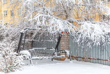 Winter cityscape on the background of a yellow brick house. The swing, enclosed by an iron fence,...