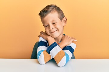 Adorable caucasian kid wearing casual clothes sitting on the table hugging oneself happy and...