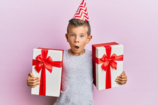 Adorable caucasian kid wearing birthday hat holding presents afraid and shocked with surprise and amazed expression, fear and excited face.