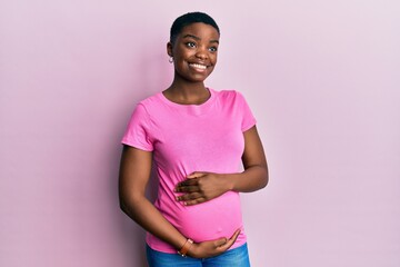 Young african american woman expecting a baby, touching pregnant belly smiling looking to the side...