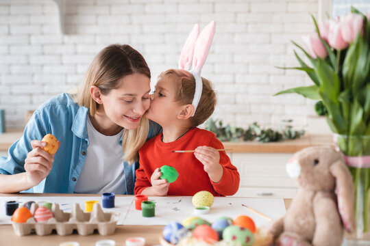 Little Cute Son Kissing His Mom While Painting Eggs For Easter In The Kitchen