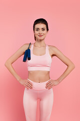 Fototapeta na wymiar Smiling sportswoman with jump rope and hands on hips isolated on pink