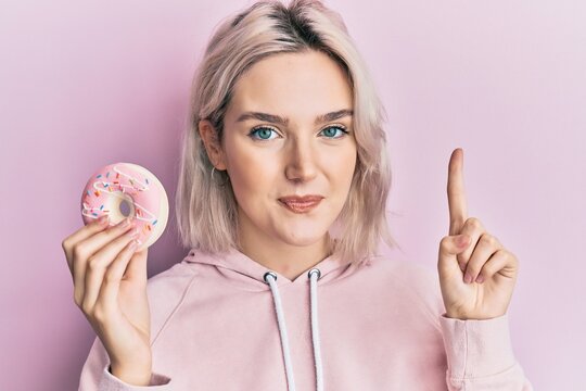 Young blonde girl holding donut smiling with an idea or question pointing finger with happy face, number one