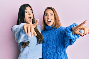 Hispanic family of mother and daughter wearing wool winter sweater pointing with finger surprised...