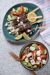 Above view of grilled lamb kofta kabobs with feta and greek salad, vertical shot on a beige stone surface