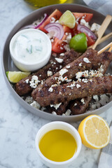 Close-up of bbq greek kofta kabobs with tzatziki sauce, feta and vegetables, vertical shot on a grey marble surface