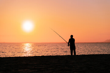 Fototapeta premium Silhouette of man fishing in waves on beach at sunset