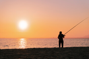 Fototapeta premium Silhouette of man fishing in waves on beach at sunset