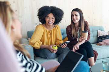 Smart entrepreneur multiethnic women talking while taking a break and showing photos with smart phone sitting on couch at coworking place.
