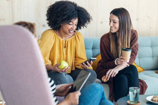 Smart entrepreneur multiethnic women talking while taking a break and showing photos with smart phone sitting on couch at coworking place.