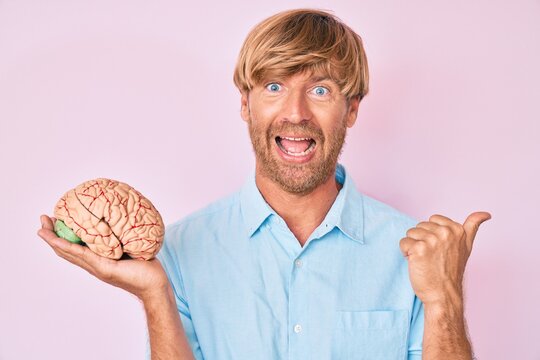 Young blond man holding brain pointing thumb up to the side smiling happy with open mouth
