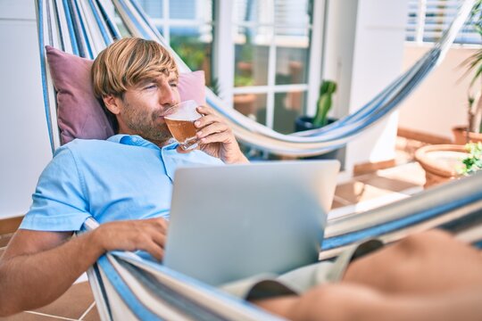 Middle age handsome man at the terrace of his house relaxing lying on a hammock working with laptop and drinking a fresh beer