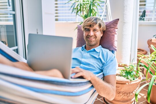 Middle age handsome man at the terrace of his house relaxing lying on a hammock working with laptop