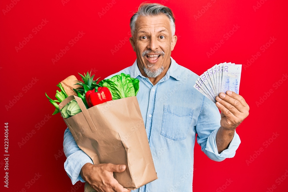 Poster middle age grey-haired man holding groceries and swedish krona banknotes smiling and laughing hard o