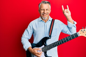 Middle age grey-haired man playing electric guitar doing rock sign smiling with a happy and cool...