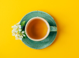 Top view of turquoise cup of green or herbal tea on saucer with spring tree flowers over bright yellow background. Springtime concept. 