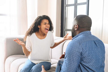 An African-American couple is quarreling sitting on the couch at home, misunderstands each other, irritated woman screaming at man, arguing emotionally. Crisis and relationship problems
