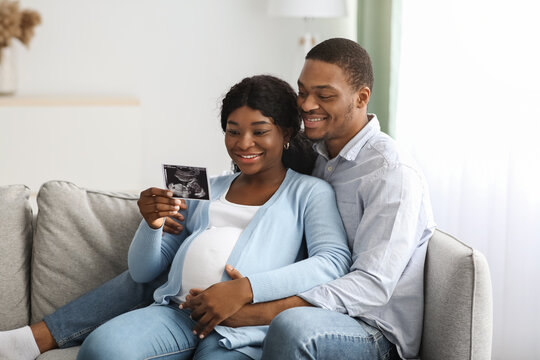 Smiling pregnant black couple holding ultrasound image, home interior