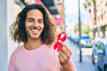 Young hispanic man smiling happy holding hiv awareness red ribbon at city. - obrazy, fototapety, plakaty