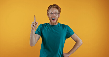 amazed redhead man in eye glasses standing with hand on hip while having idea isolated on yellow