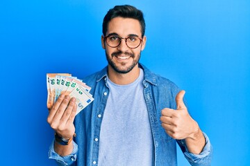 Young hispanic man holding euro banknotes smiling happy and positive, thumb up doing excellent and approval sign - obrazy, fototapety, plakaty