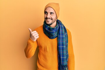 Young hispanic man wearing wool sweater and winter scarf pointing thumb up to the side smiling...