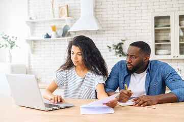 Concentrated African-American couple sits at the kitchen table with a laptop at home, calculating finance, checking bank account balance, a biracial woman looks through papers, a man takes a notes - Powered by Adobe