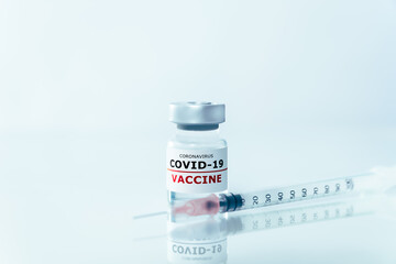Covid-19 vaccine glass vial on the white surface with a plain white background and a syringe laying in front of the glass vial.