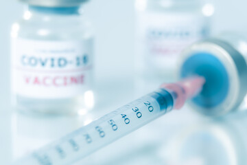 Close up of the syringe inserted in the coronavirus vaccine vial layiong on the white shiny surface with a 2 blurred covid vaccine vials on the background