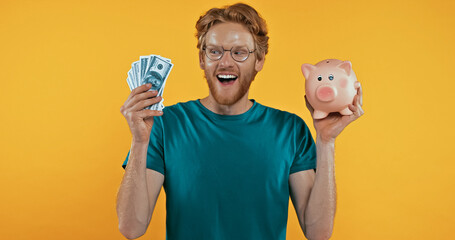 happy redhead man holding piggy bank and cash isolated on yellow