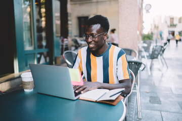 Happy male user in optical spectacles installing application for creating blog publication via laptop technology, smiling hipster guy searching content information while browsing wireless on netbook