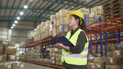 young asian chinese woman pointing finger and checking list in documents on clipboard while working at large warehouse.