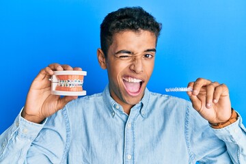 Young handsome african american man holding invisible aligner orthodontic and braces winking...